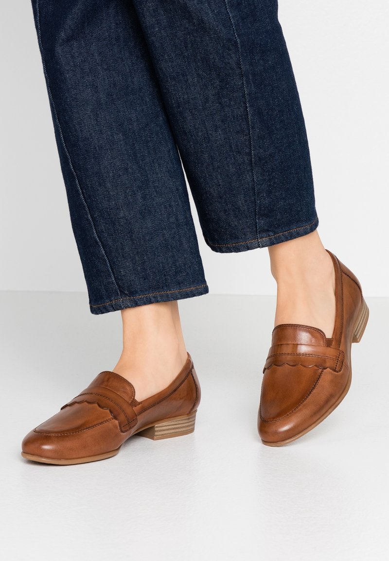 Personne portant des mocassins en cuir marron à talon bas avec un détail en bord festonné et un jean en denim bleu foncé coupé sur un sol clair.