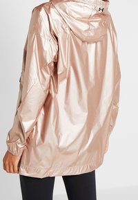 Lekka, metaliczna kurtka przeciwwiatrowa w kolorze różowego złota z kapturem, elastycznymi mankietami i lekko błyszczącą teksturą. Cechuje ją sportowy design.
