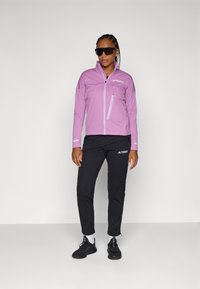Lichtpaarse lichtgewicht jas met een ritszak en reflecterende accenten, gecombineerd met zwarte broek met een logo en zwarte sporty schoenen.