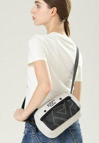 Borsa a tracolla con un corpo in tessuto grigio e una tasca frontale nera, decorata con bold testo bianco e design geometrici. Includa una tracolla regolabile.