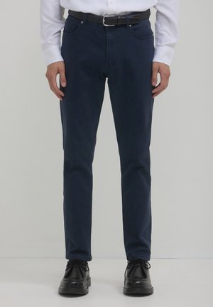 Mann in dunkelblauben Slim-Fit-Hosen, schwarzem Gürtel, schwarzen Schnürschuhen und einem weißen Langarmhemd, der vor einem schlichten Hintergrund steht.