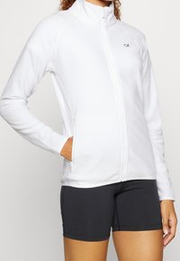 Bílá fleecová bunda s vysokým límcem a předním zipem, vybavená bočními zipovými kapsami, navržená pro přiléhavý vzhled a vrstevnatý styl.