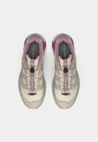 Μπεζ και μοβ αθλητικά παπούτσια με υφασμένο άνω μέρος, σύστημα κορδονιών και μαλακή σόλα. Σημαντικά στοιχεία περιλαμβάνουν μια θηλιά έλξης και διακοσμήσεις με το λογότυπο.