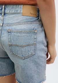 Ανοιχτό μπλε σορτς από τζιν με ξέφτισμα στο τελείωμα, με τσέπες στο πίσω μέρος και ταμπέλα από καφέ δέρμα στη μέση. Ραφές σε αντίθετο χρώμα.