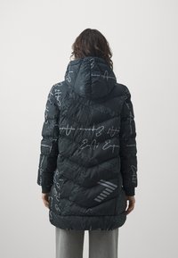 Kvinna iklädd en svart huva pufferjacka med grå grafisk text och symboler, stående med ryggen mot kameran framför en enfärgad bakgrund.