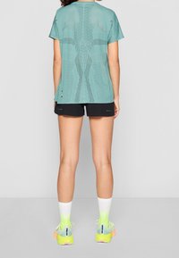 Kvinna som står med ryggen vänd, iklädd en teal perforerad kortärmad topp, svarta shorts, vita och neongula strumpor samt neongula löparskor.