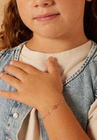 Goldkette mit Armbändern in Form von rosa Schmetterlingen, die am Handgelenk eines Kindes getragen wird, vor einem Hintergrund aus einer hellfarbigen Jeansjacke.