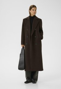 Manteau en laine marron à double boutonnage avec de grands boutons noirs, poches latérales et coupe longue, associé à un sac noir et un pantalon en cuir.