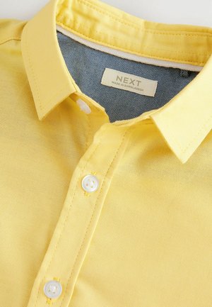 Chemise jaune à boutons, avec une texture douce, dotée d'un col et de boutons blancs, associée à une doublure en denim au niveau du cou.