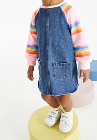 Denim jurk met een roze en gestreept longsleeve shirt. Heeft twee steekzakken aan de voorkant en wordt aangevuld met witte klittenbandschoenen.