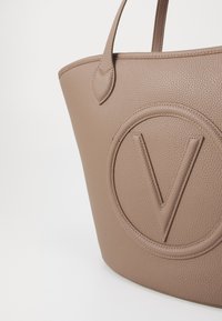 Borsa tote in pelle beige con texture, dotata di un logo circolare in rilievo che presenta una 'V' al centro. Manici doppi attaccati ai lati.