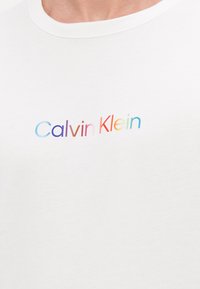 Hvit bomulls T-skjorte med et flerfarget "Calvin Klein"-logo i en moderne, avrundet skrifttype sentrert på brystet.