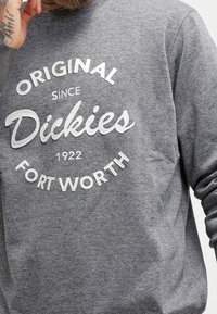 Felpa grigia con testo bianco che recita "Originale dal 1922 Dickies Fort Worth", indossata da una persona con la mano tatuata vicino al viso.