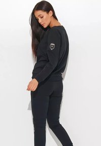 Czarna bluza z miękkiego materiału, z okrągłym dekoltem i długimi rękawami. Na lewym rękawie znajduje się ozdobna naszywka z napisem "nu". Połączona z pasującymi spodniami.