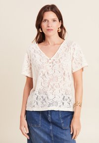 Top in pizzo color crema con maniche corte, scollatura a V profonda e tre bottoni sul davanti; presenta un motivo floreale e una finitura testurizzata.