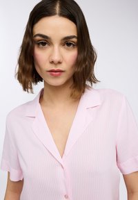 Camicia button-up a maniche corte di colore rosa chiaro con fini strisce bianche verticali, colletto a revers e vestibilità rilassata. Tessuto dalla superficie liscia.