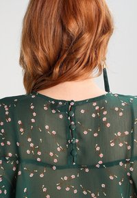 Blouse vert foncé à imprimé floral, tissu transparent, col rond, et fermeture au dos avec quatre boutons recouverts de tissu.