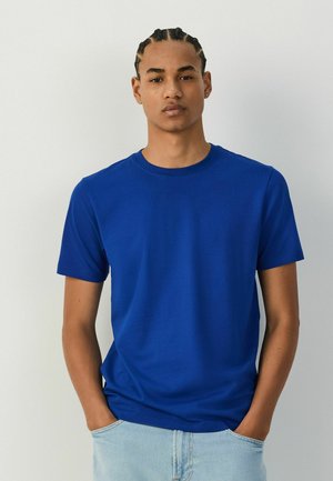 Ung mand med flettet hår iført en ensfarvet blå t-shirt med korte ærmer og lyseblå jeans, stående med hænderne i lommerne.
