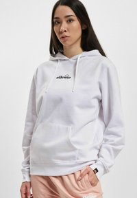 Ungdommelig kvinne iført en hvit ellesse hettegenser og ferskenfargede ellesse joggebukser, med en hånd i lommen, mot en enkel lys bakgrunn.