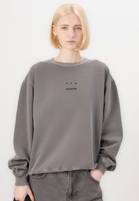 Sweatshirt grigio realizzato in morbido tessuto, con un colletto a girocollo a costine, spalle scese e un logo nero centrato sul petto.