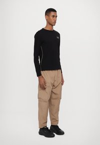 Camisola de manga longa preta com logotipo dourado e piping lateral, combinada com calças cargo bege com bolsos laterais e um acabamento texturizado. Ténis pretos.