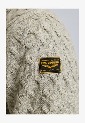 Beżowy sweter dziany z wzorem w warkocze, z czarną i żółtą metką marki "PME LEGEND" przyszytą na piersi.