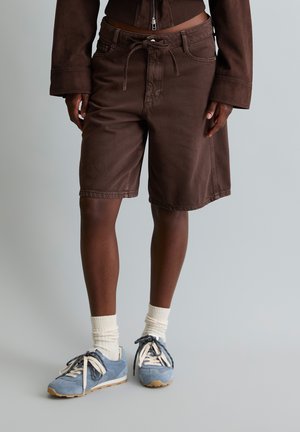 Personne portant un short marron jusqu’aux genoux avec un cordon, une veste marron, des chaussettes blanches et des baskets bleues, debout sur un sol gris.