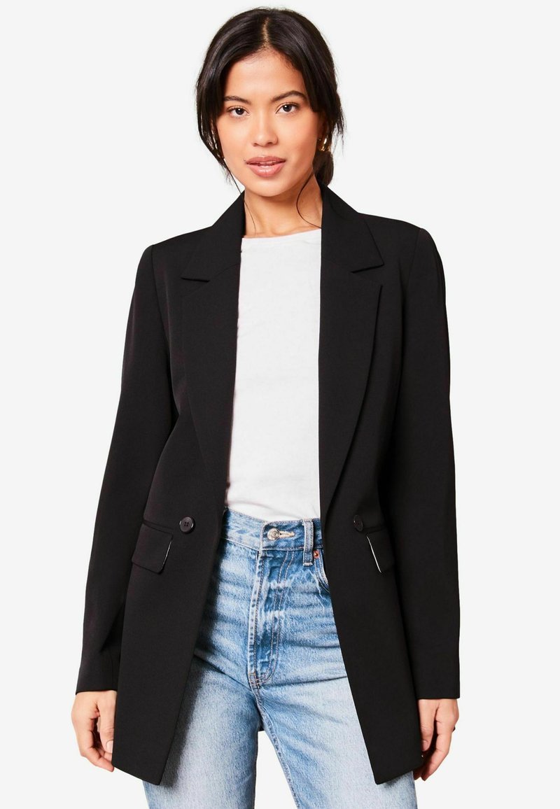 Blazer negro con un diseño ajustado, solapas muestradas, dos botones y bolsillos laterales, llevado sobre una blusa blanca con jeans de cintura alta en azul claro.