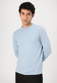 Himmelblauer Langarm-Pullover mit Rundhalsausschnitt aus weichem Stoff. Mit einem gerippten Kragen und klassischem Schnitt. Getragen mit schwarzen Hosen.