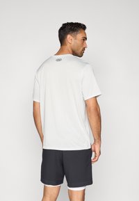 Camiseta deportiva blanca con cuello redondo, mangas cortas y un logo negro en la parte trasera. Combinada con pantalones cortos gris oscuro y pantalones cortos interiores blancos.