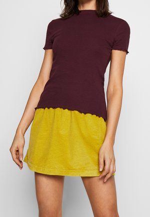 Kobieta w dopasowanym, ciemnofioletowym topie z krótkim rękawem i żółtej, sztruksowej mini spódnicy z wysokim stanem na jednolitym tle.