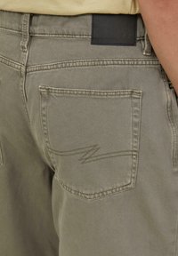 Spodnie khaki widok z tyłu, z skórzaną metką w talii, tylną kieszenią ze zygzakowym szyciem oraz gładką denimową fakturą.