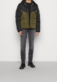 Oppustelig jakke med en sort øverste del og olivengrønne nederste del. Har hætte, lynlåslommer og quiltet tekstur. Parret med grå jeans og sorte sko.