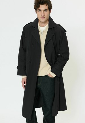 Ung mand iført en lang sort trenchcoat over en beige trøje med lynlås og mørke jeans, stående med den ene hånd i lommen.