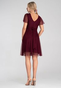 Bordeauxfarbiges Kleid mit kurzen, transparenten Ärmeln, einem V-Ausschnitt im Rücken und einer gebundenen Taille. Halbtransparenter Rock sorgt für Volumen; kombiniert mit Riemchensandalen.