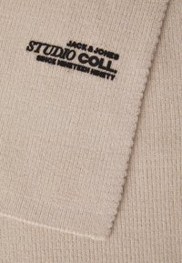 Beige gebreide sjaal met ribbelstructuur. Heeft zwarte geborduurde branding "JACK & JONES STUDIO COLL. SINCE NINETEEN NINETY."