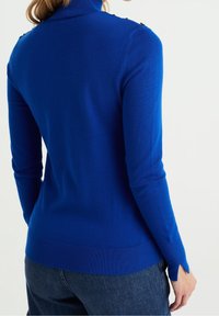 Blå, ribbad polotröja med långa ärmar, accentuerad med knapppar på axlarna och skärningar vid ärmarnas manschetter.