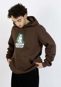 Ruskea huppari, jossa on Muumi-hahmon kuvio ja teksti "MOOMIN HIKING CLUB." Siinä on kengurutasku ja kiristysnyörillä varustettu huppu.