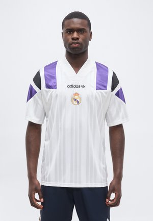 Młody mężczyzna w białej koszulce piłkarskiej Adidas Real Madrid z fioletowymi i czarnymi akcentami, stojący na jasnym, jednolitym tle.