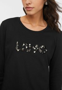Top nero a maniche lunghe con scollo rotondo. Presenta un motivo "LIU JO" realizzato con paillettes nere lucide e argento. Tessuto morbido.