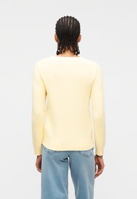 Personne avec des cheveux tressés portant un pull à manches longues jaune clair et un jean bleu clair, vue de dos sur un fond blanc.