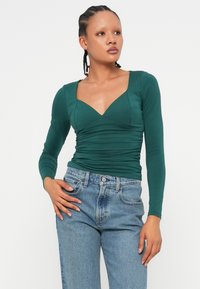 Πράσινη μπλούζα μακρυά μανίκια με βαθύ V-ντεκολτέ και πτυχώσεις, συνδυασμένη με ανοιχτό μπλε τζιν. Το ύφασμα φαίνεται μαλακό και εφαρμοστό.