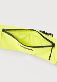 Φωτεινή κίτρινη τσάντα μέσης με μαύρο φερμουάρ μισάνοιχτο, εμφανίζοντας το κενό εσωτερικό και το μαύρο λογότυπο HOKA στο μπροστινό μέρος.