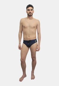 Costume da bagno slip blu navy con coulisse frontale, tessuto leggero e piccolo logo decorativo sul davanti. Modello in piedi a piedi nudi.