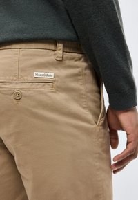 Pantalon beige avec poche arrière et étiquette Marc O’Polo, porté avec une chemise à manches longues gris foncé, main relaxée sur le côté.