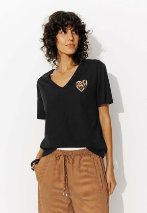 Femme aux cheveux bouclés portant un T-shirt noir à col en V avec un design de cœur "More Amor" et un pantalon rayé marron, se tenant devant un fond blanc.