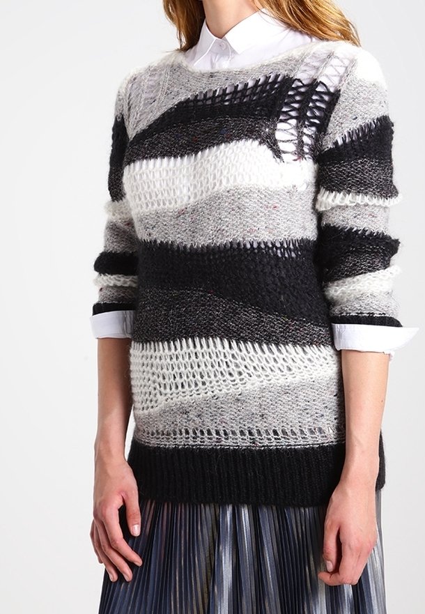 Pull en maille noir, gris et blanc avec diverses textures ; présentant des mailles ouvertes et des sections unies. Porté par-dessus une jupe plissée.