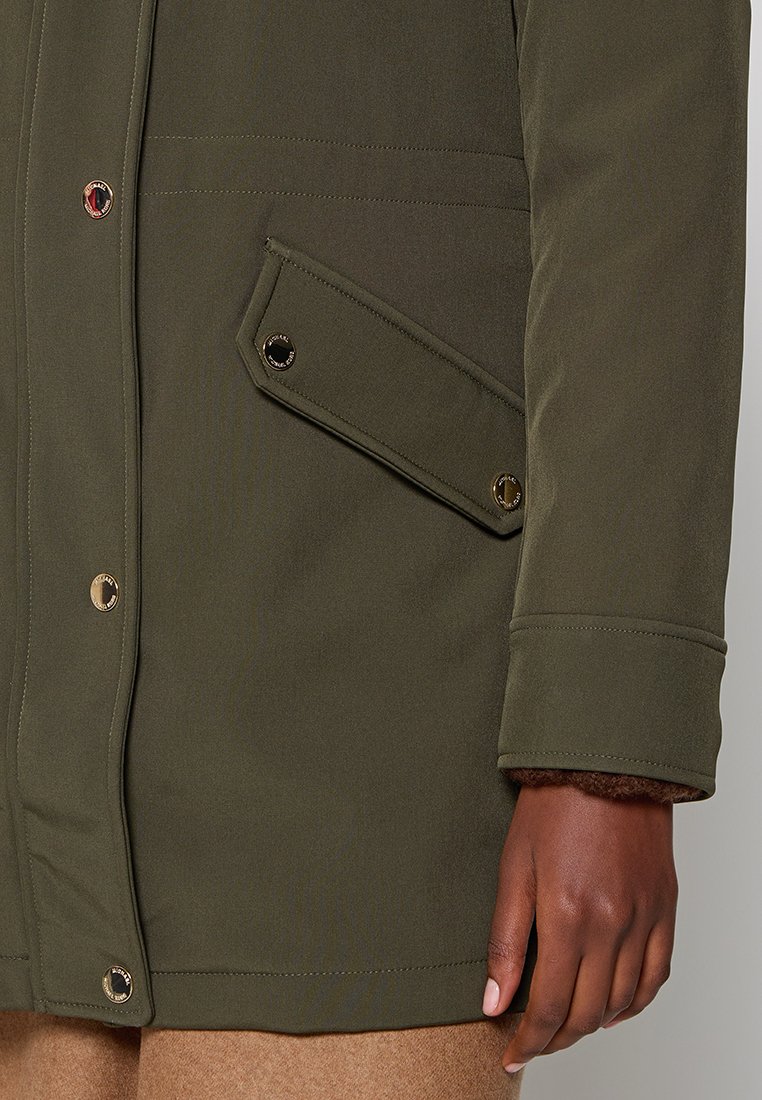 Giacca esterna verde con accessori dorati, caratterizzata da una tasca sul petto e una morbida fodera marrone testurizzata sul polsino.