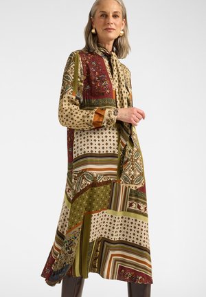 Frau mit grauen Haaren, die ein langes Patchwork-Kleid mit floralen, gestreiften und geometrischen Mustern in Beige-, Grün-, Braun- und Rottönen trägt.