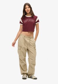 Bordeaux cropped t-shirt met "SUPERDRY" logo, witte gestreepte mouwen; beige cargo pants met zijzakken, trekkoord in de taille, losse pasvorm.